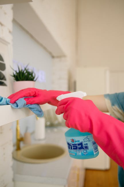 Close-up of a person wearing pink rubber gloves holding a spray bottle of cleaning product, applying sanitiser to a white kitchen shelf. A blue cleaning cloth is draped over the shelf edge, which also supports a small potted green plant. The background features a blurred view of a kitchen with a beige countertop and a sink, showcasing smooth surfaces and a clean, well-maintained environment. The scene reflects domestic surface cleaning and sanitisation, aligning with Highbury Cleaner's focus on professional cleaning services for residential spaces, as indicated on their highburycleaner.co.uk webpage about avoiding hidden charges in cleaning quotes.