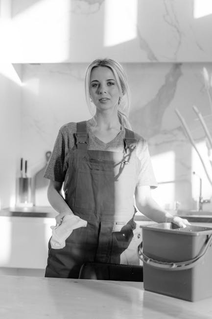 A young woman with light-colored hair tied back, wearing a short-sleeved t-shirt and an apron, is standing in a modern kitchen. She is holding a cloth in her right hand and resting her left hand on a bucket or cleaning container placed on the countertop. The kitchen features a marble backsplash, stainless steel sink, and sleek countertops, with natural light illuminating the space. The surfaces appear clean and well-maintained, reflecting a professional approach to domestic cleaning and sanitisation, consistent with Highbury Cleaner’s emphasis on offering transparent and comprehensive cleaning quotes.