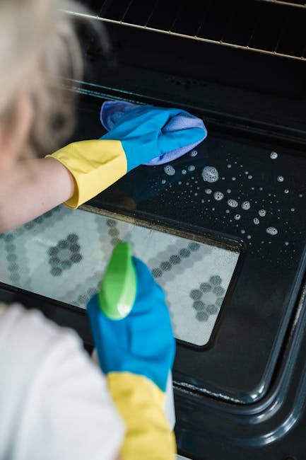 A person wearing yellow and blue cleaning gloves is cleaning the glass surface of an oven door, which is part of a black oven with a visible interior. The oven's glass shows some soap bubbles and streaks indicating recent cleaning. The scene is well-lit, with a focus on the surface cleaning process. This image exemplifies the thorough approach taken by Highbury Cleaner to maintain hygiene and cleanliness in domestic kitchen environments, specifically during oven cleaning tasks as highlighted in the page titled 'Common problems when booking Highbury oven cleaning' on highburycleaner.co.uk.