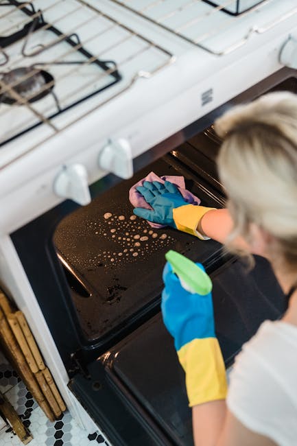A person wearing blue rubber gloves and yellow sleeves is cleaning the interior of a black oven with a pink cleaning cloth. The oven is situated below a white gas stove with visible burners and control knobs, in a kitchen with black and white checkered tile flooring. The oven door is open, revealing the dark interior, and the person is focused on surface cleaning to maintain hygiene. The surrounding kitchen surfaces and floor appear clean and well-maintained. This image exemplifies professional domestic cleaning services offered by Highbury Cleaner, emphasizing detailed oven sanitisation and deep cleaning processes.