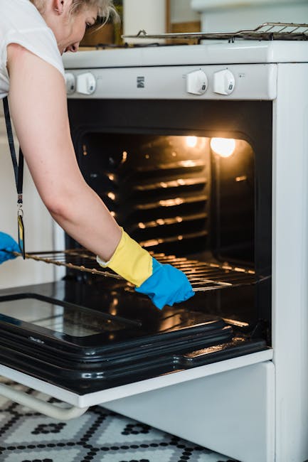 A person wearing yellow and blue cleaning gloves is cleaning the glass surface of an oven door, which is part of a black oven with a visible interior. The oven's glass shows some soap bubbles and streaks indicating recent cleaning. The scene is well-lit, with a focus on the surface cleaning process. This image exemplifies the thorough approach taken by Highbury Cleaner to maintain hygiene and cleanliness in domestic kitchen environments, specifically during oven cleaning tasks as highlighted in the page titled 'Common problems when booking Highbury oven cleaning' on highburycleaner.co.uk.