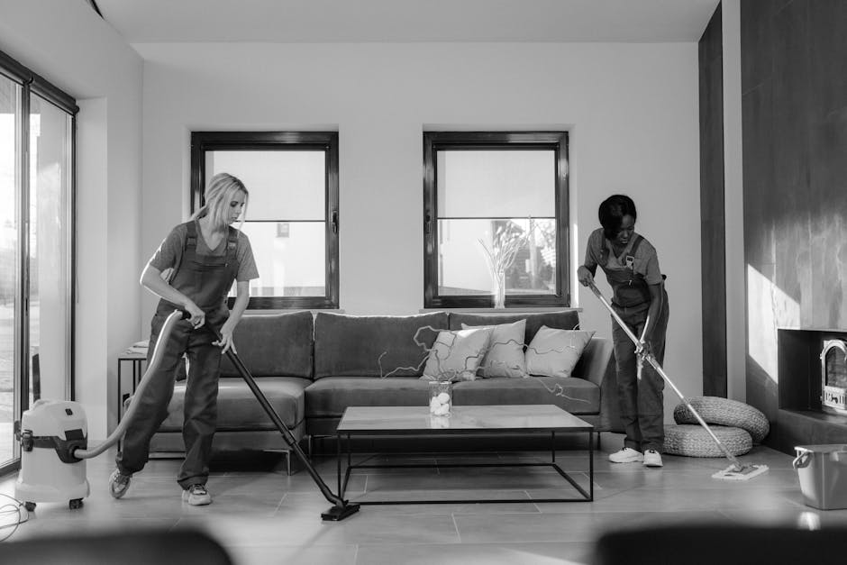 Two professional cleaners in an open-plan living room are engaged in surface cleaning and deep cleaning activities. One cleaner, on the left, is using a vacuum cleaner to thoroughly clean the wooden floor, while the other, on the right, is using a mop on the tiled area near the fireplace. The room features a modern, minimalist design with a dark-colored sofa adorned with cushions, positioned against the wall with two large windows bringing in natural light. On the coffee table in front of the sofa, there is a small decorative object. The room appears well-maintained and spotless, with gleaming surfaces and no visible dust or dirt. Visible cleaning tools and equipment, including the vacuum and mop, suggest a thorough sanitisation process, consistent with high-quality domestic cleaning standards provided by Highbury Cleaner for end of tenancy services.