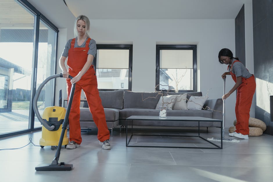 Two professional cleaners in an open-plan living room are engaged in surface cleaning and deep cleaning activities. One cleaner, on the left, is using a vacuum cleaner to thoroughly clean the wooden floor, while the other, on the right, is using a mop on the tiled area near the fireplace. The room features a modern, minimalist design with a dark-colored sofa adorned with cushions, positioned against the wall with two large windows bringing in natural light. On the coffee table in front of the sofa, there is a small decorative object. The room appears well-maintained and spotless, with gleaming surfaces and no visible dust or dirt. Visible cleaning tools and equipment, including the vacuum and mop, suggest a thorough sanitisation process, consistent with high-quality domestic cleaning standards provided by Highbury Cleaner for end of tenancy services.