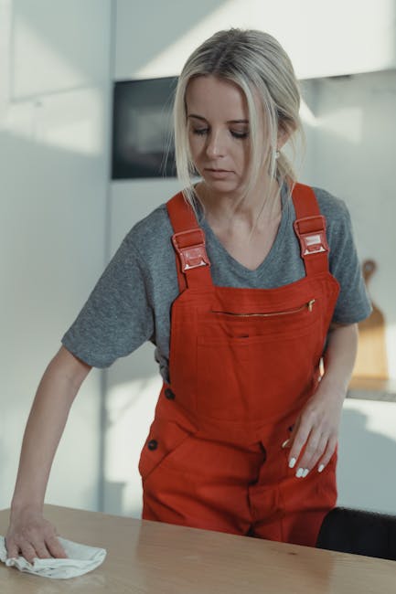 A female cleaner with blonde hair, dressed in a grey t-shirt and red overalls, is engaged in surface cleaning in a domestic kitchen setting. She is wiping a wooden table with a white cloth, surrounded by natural light, with a modern, minimalist kitchen background featuring white and grey cabinetry. The surface appears clean and polished, reflecting the ambient light, indicating effective deep cleaning and maintenance. Visible cleaning tools include the cloth she is using, and no other tools are apparent. The environment looks well-organized and hygienic, aligning with professional cleaning standards promoted by Highbury Cleaner in their house cleaning guide and local cleaning services.
