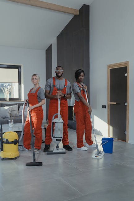 Three professional cleaners from Highbury Cleaner, dressed in orange overalls and grey shirts, stand in a spacious, well-lit modern living room with high ceilings. They are equipped with cleaning tools: one holding a vacuum cleaner, another with a mop and bucket, and the third with a cleaning cloth and spray. The room features a polished grey tiled floor, a light-colored wall, and a large window allowing natural light to illuminate the area, highlighting the spotless and shiny surface. A comfortable sofa with cushions and a small glass table are visible in the background, indicating a tidy and maintained domestic space. The scene emphasizes surface cleaning, deep cleaning, and maintenance services provided by Highbury Cleaner to ensure hygiene and cleanliness in residential environments.