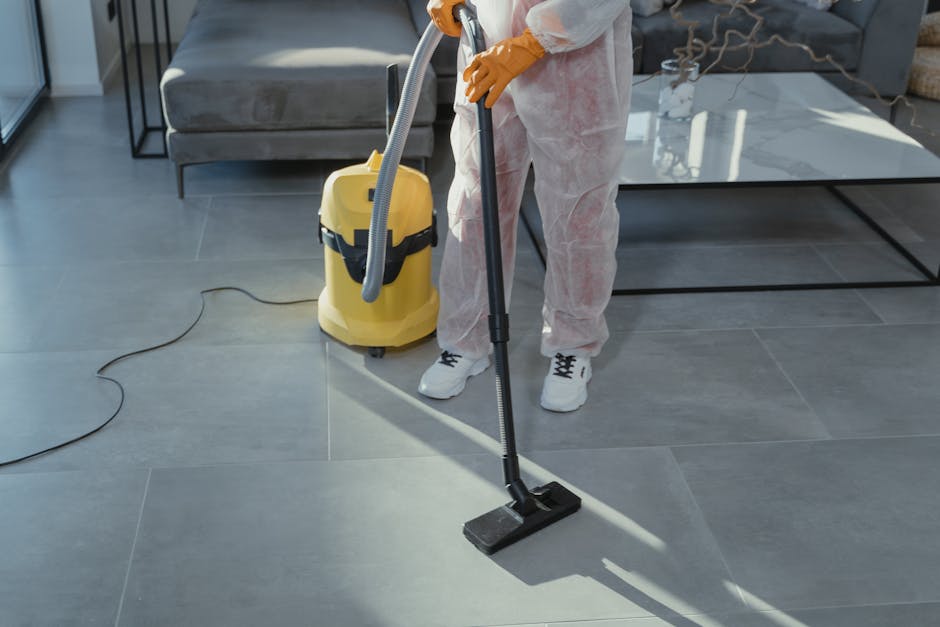 A female cleaner with blonde hair, dressed in a grey t-shirt and red overalls, is engaged in surface cleaning in a domestic kitchen setting. She is wiping a wooden table with a white cloth, surrounded by natural light, with a modern, minimalist kitchen background featuring white and grey cabinetry. The surface appears clean and polished, reflecting the ambient light, indicating effective deep cleaning and maintenance. Visible cleaning tools include the cloth she is using, and no other tools are apparent. The environment looks well-organized and hygienic, aligning with professional cleaning standards promoted by Highbury Cleaner in their house cleaning guide and local cleaning services.