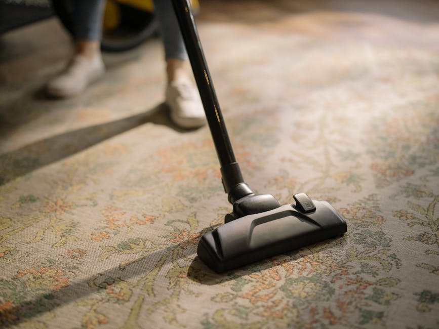 Close-up of a person using a vacuum cleaner on a patterned carpet in a residential living room, with a black vacuum head and a long metal wand. The carpet features intricate floral designs in muted shades of green, orange, and beige. Part of a person's leg in dark pants and white shoes is visible in the background, standing on the carpet. The lighting is natural and soft, highlighting the cleanliness of the carpet and the vacuuming process. This image illustrates surface cleaning and maintenance typical of professional domestic cleaning services provided by Highbury Cleaner near Highbury N5.