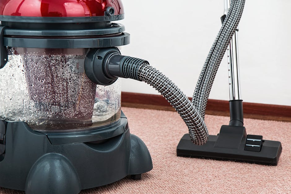 A professional cleaning technician dressed in full protective gear, including a white disposable suit, mask, and gloves, is performing deep cleaning on a beige carpet in a well-lit, modern living room. The technician is using a specialized steam cleaning machine, with steam and mist visible on the carpet surface. The room features contemporary décor with wooden shelving units, potted plants, and framed artwork on the walls, and appears clean and well-maintained. The scene emphasizes thorough surface cleaning and sanitisation, performed by Highbury Cleaner as part of their residential cleaning services, highlighting attention to detail in hygiene and stain removal.