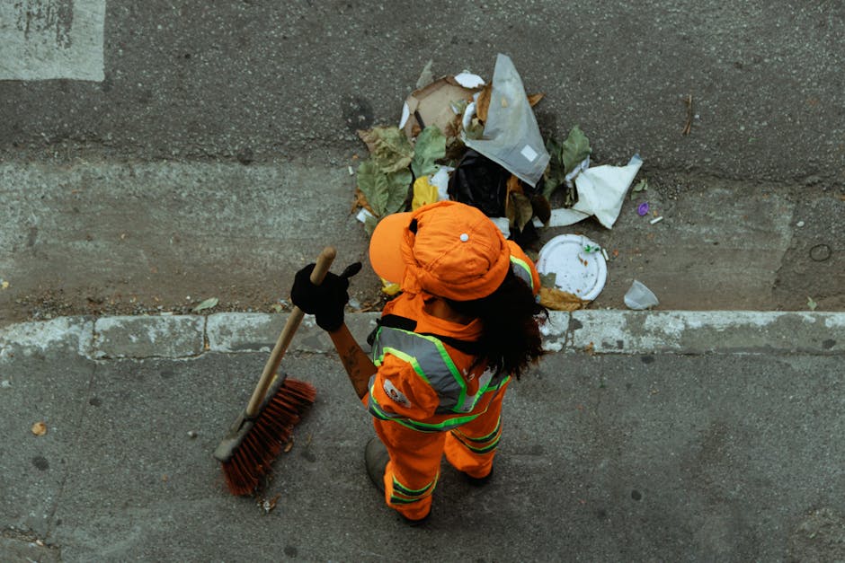 A street cleaner dressed in bright orange high-visibility clothing with reflective stripes is sweeping the pavement with a long-handled broom next to a green waste collection bin covered with a black 