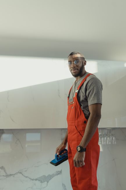 A professional worker performing high-rise exterior glass surface cleaning on a modern building balcony. The worker, wearing a safety helmet, harness, and high-visibility vest, is seated on the balcon
